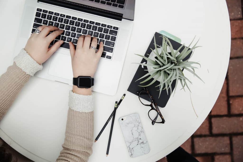 A woman is using a laptop on a round, white table. The photo is taken from above, so we can only see her arms as she types on the computer. We also see her workspace, which is minimalistic and clean. On the table around her she has two black pencils, her iPhone, a pair of glasses, a small black planner, and a green plant.