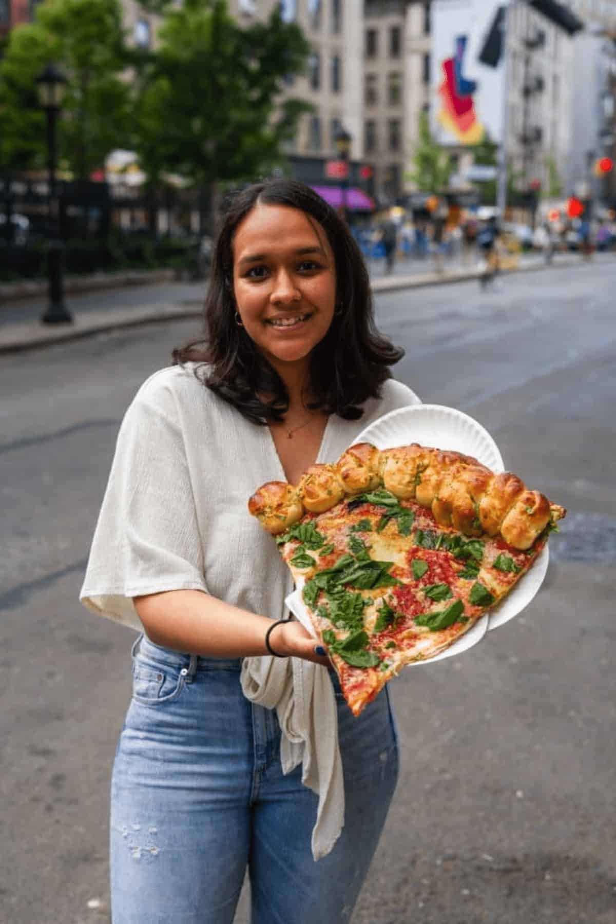 woman standing on sidewalk holding up a large piece of pizza