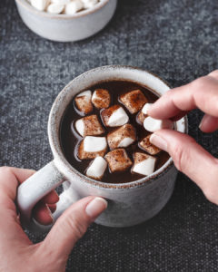 someone holding a coffee cup of hot carob and putting more marshmallows on top