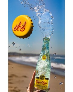 phoo of a topo chico spraying in front of the ocean