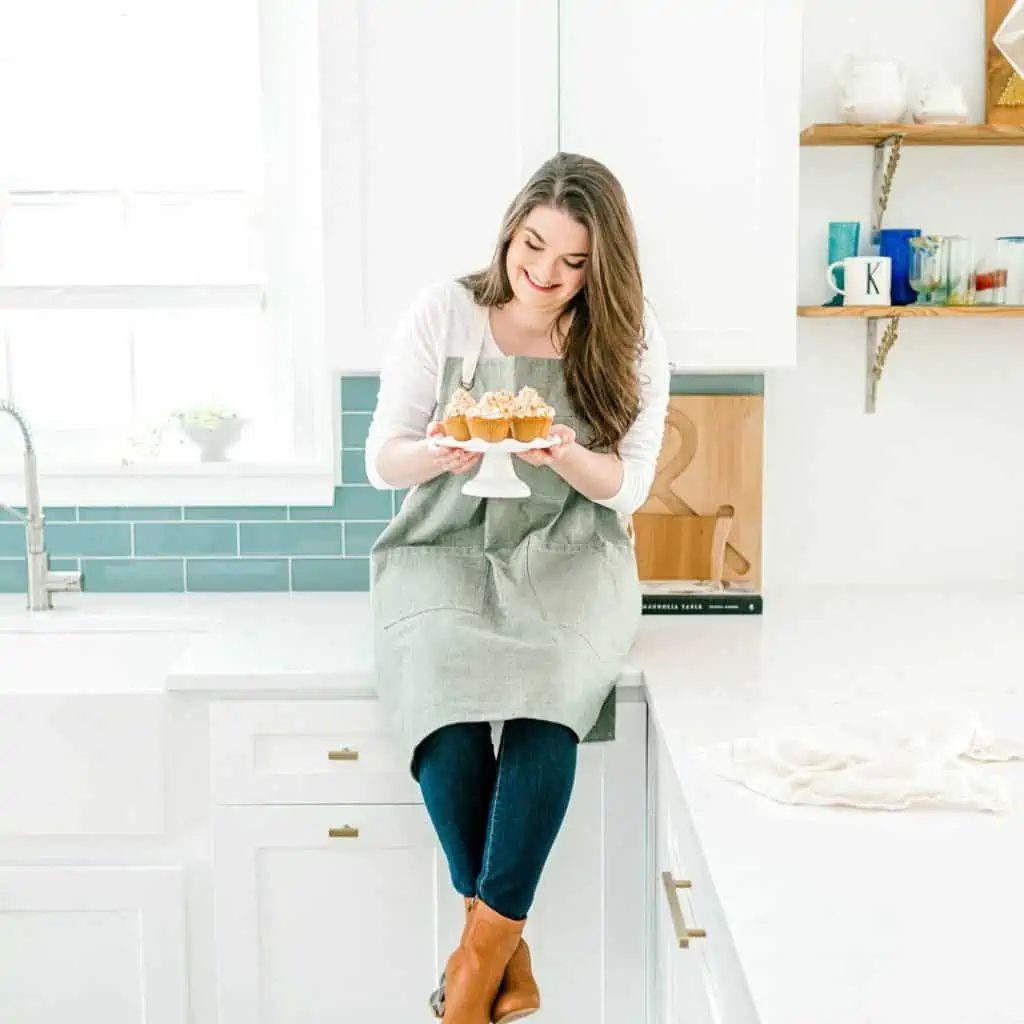woman sitting on a counter smiling at a cake stand of cupcakes