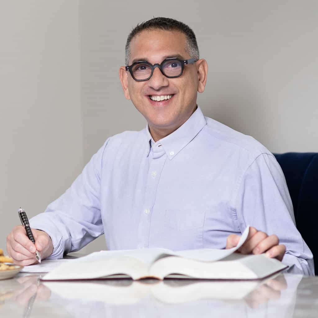 Man with black glasses and purple shirt smiling and writing on the pages of an open book