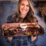 Susie Bulloch smiling while holding a large tray of sliced smoked brisket with a small bowl of barbecue sauce in the center. She is wearing a plaid shirt and standing in front of a smoker, with visible steam rising behind her.