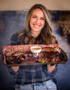 Susie Bulloch smiling while holding a large tray of sliced smoked brisket with a small bowl of barbecue sauce in the center. She is wearing a plaid shirt and standing in front of a smoker, with visible steam rising behind her.