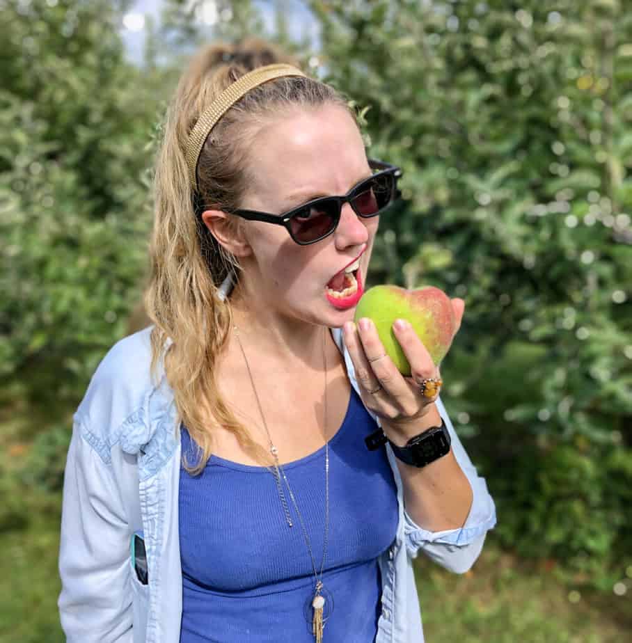 blonde woman with a blue shirt and glasses about to take a bite of a green apple