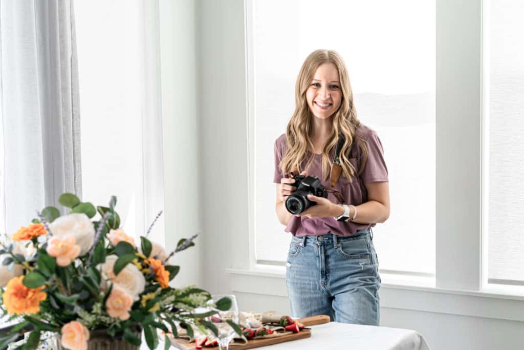 woman with blonde hair holding a camera and smiling behind a bouquet of flowers and charcuterie board