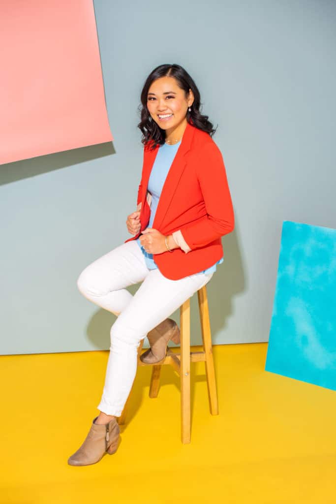 woman sitting on a wooden stool in white pants, blue shirt, and red blazer