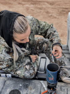 woman with blonde hair and camouflage outfit pouring packets into a Jetboil camping stove