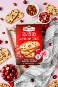 box of THINaddictives cranberry almond thin cookies surrounded by almonds, cranberries, and cookies