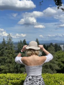 woman with blonde hair, a white top, and cream colored hat back facing the camera outdoors with trees and clouds