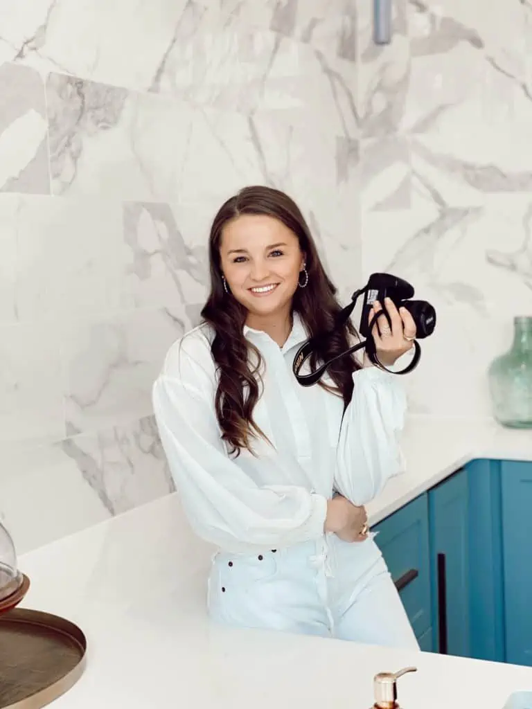 woman with white shirt, blue jeans, and brown hair holding a camera and smiling