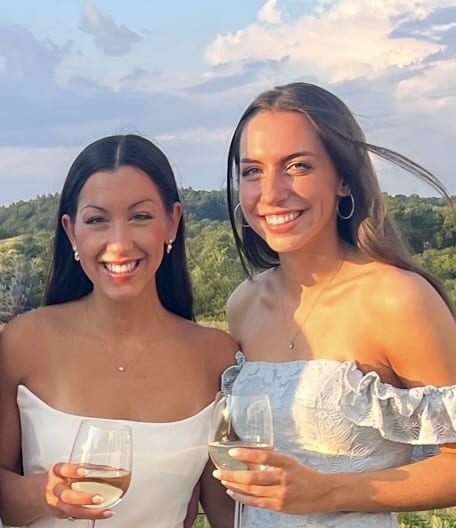 two women with brown hair in dresses, smiling, and holding wine glasses outdoors