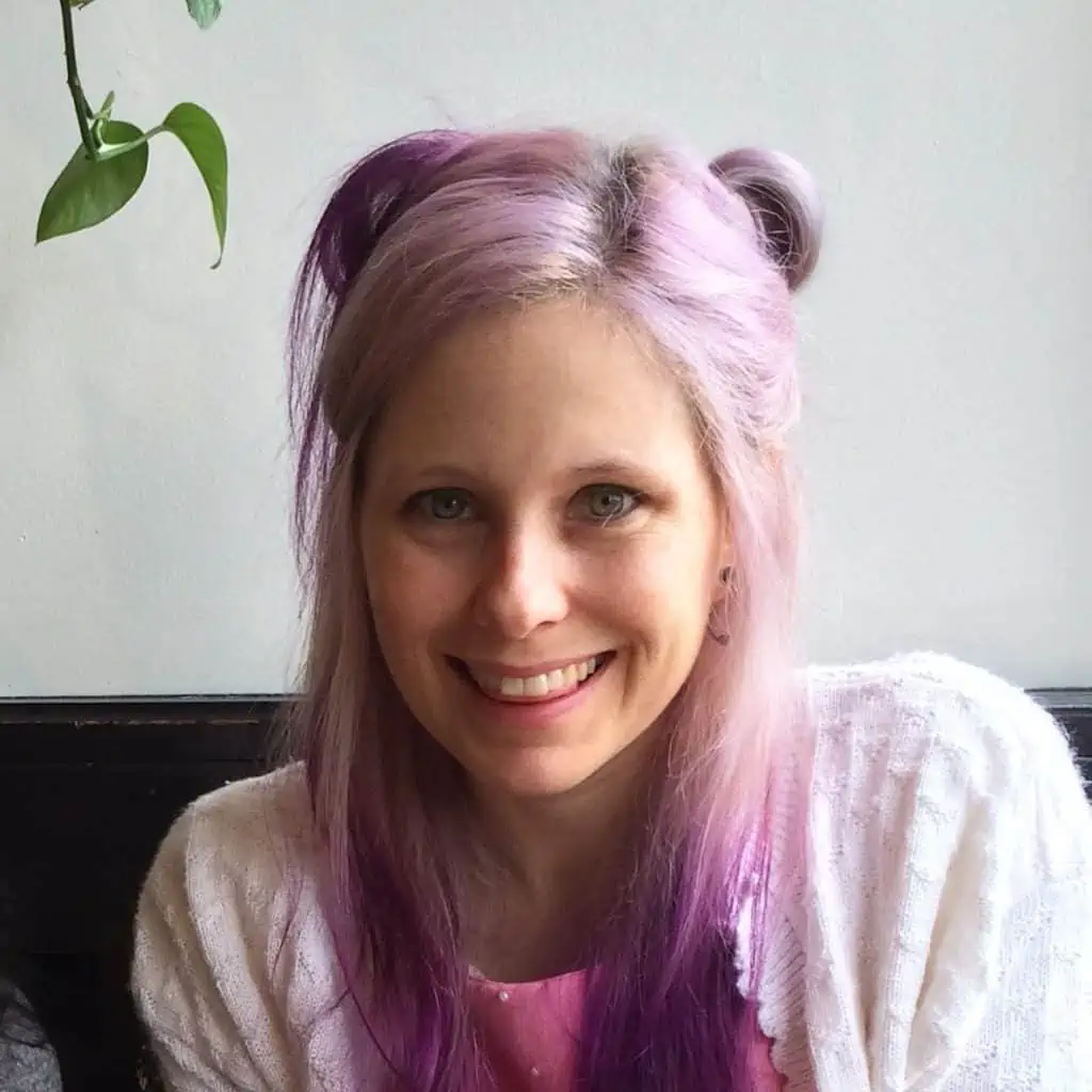 woman with purple hair, pink shirt, and white cardigan smiling