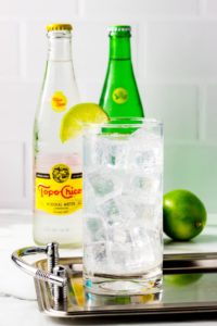 texas ranch water in a glass cup with ice and a slice of lime on top in front of TopoChico bottle and green bottle