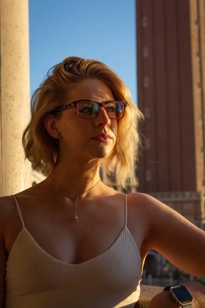 woman with blonde short hair, a cream colored tank top, and glasses looking off to the side