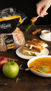 hand drizzling honey over a sandwich next to a loaf of bread, two apples, and soup