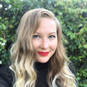 woman with blonde hair wearing a black sweater smiling in front of greenery