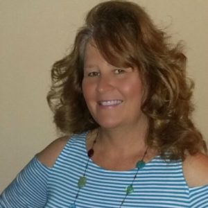 woman with brown hair wearing a blue shirt and necklace smiling