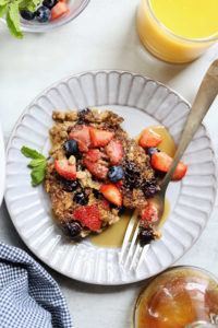 baked oatmeal with strawberries and blueberries on a plate next to a bottle of syrup and cup of orange juice