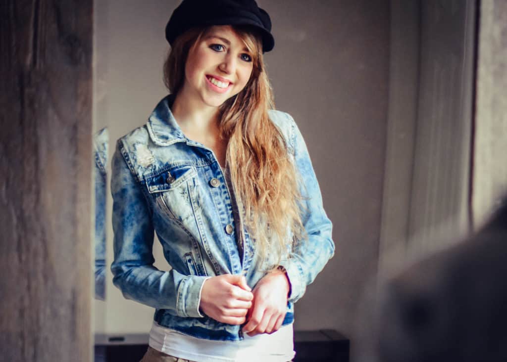 blonde woman wearing a jean jacket and black hat standing in front of a mirror and smiling