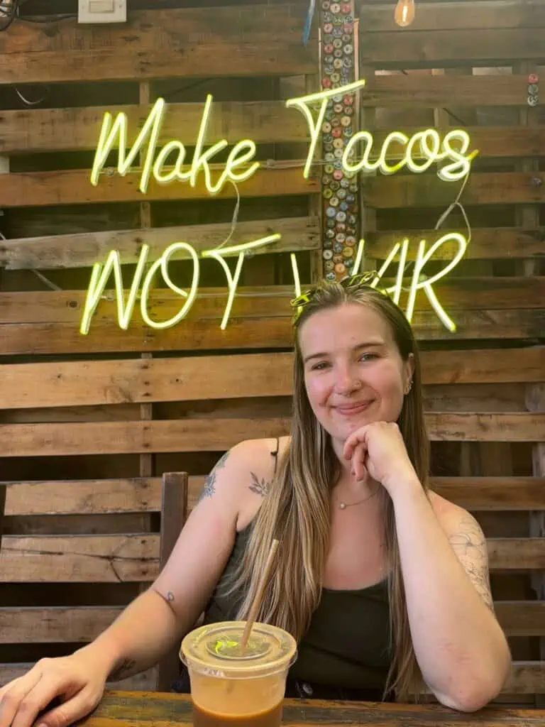 blonde woman posing in front of a drink and neon yellow sign reading make tacos