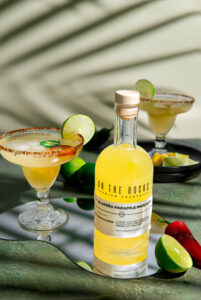 margarita with a slice of lime next to a yellow bottle of On of the Rocks jalapeño pineapple margarita and a lime and chile
