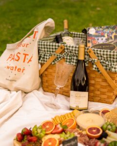 picnic with a charcuterie board filled with fruits and crackers next to a picnic basket, wine bottle, wine glass, and tote