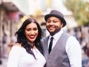 woman with dark hair wearing a white long sleeve smiling next to a man wearing a tie, vest, and gray hat