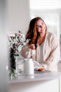 woman with dark hair wearing a white cardigan smiling off to the side of the camera and pouring flour into a bowl