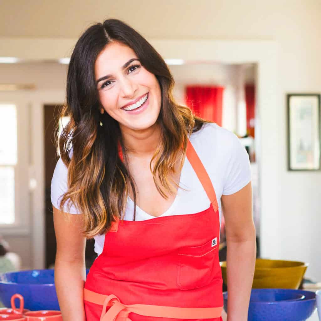 A smiling woman with long, dark brown hair wears a white shirt and a bright red apron. She is leaning slightly forward in a well-lit kitchen. The background shows colorful bowls and kitchenware, creating a cheerful and welcoming atmosphere.