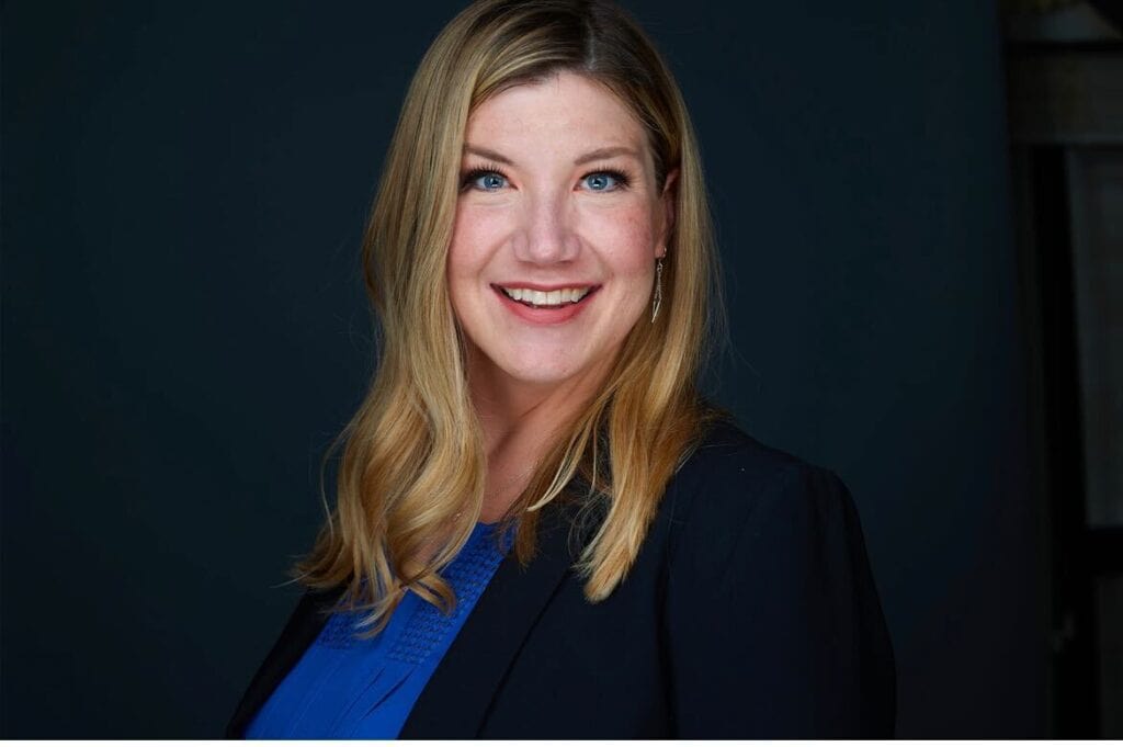 a professional portrait of a smiling woman with long, wavy blonde hair. She is wearing a blue blouse and a black blazer, with minimal jewelry including dangling earrings.