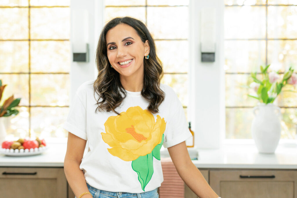 Yumna Jawad smiling warmly in a bright kitchen, wearing a white shirt with yellow design and turquoise earrings.
