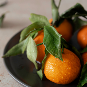 Close-up of fresh, vibrant oranges with leaves, with water droplets visible on the surface, arranged on a dark plate against a light-colored tabletop, conveying a sense of freshness and natural taste.