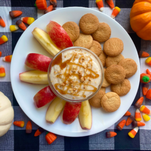 A festive autumn snack plate featuring apple slices, a glass of caramel-drizzled yogurt, and vanilla cookies on a white plate, surrounded by scattered candy corn and a small pumpkin, on a checkered tablecloth.