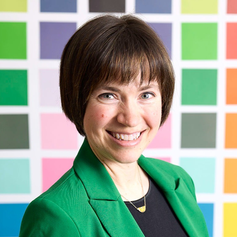 A smiling woman with short dark hair and a bright green blazer stands in front of a colorful wall of squares in various shades of green, blue, pink, orange, and gray. Her cheerful expression and the vibrant background suggest creativity and positivity.