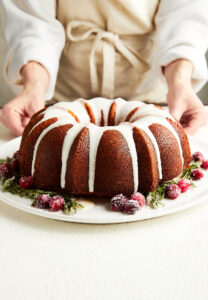 A bundt cake with strawberries around the edges and icing dripping down the sides in the foreground, a woman with a white shirt and tan apron in the background.