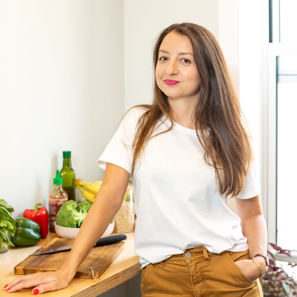 A woman with long brown hair and pink lipstick wears a white t-shirt and brown pants. She stands in front of a counter top with a wooden cutting board.