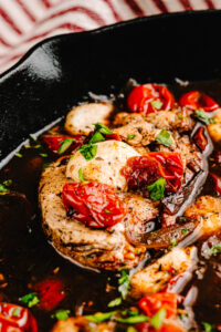 A skillet containing balsamic chicken with roasted cherry tomatoes, topped with fresh basil. The chicken appears golden-brown and seasoned, with balsamic glaze and juices visible in the pan.