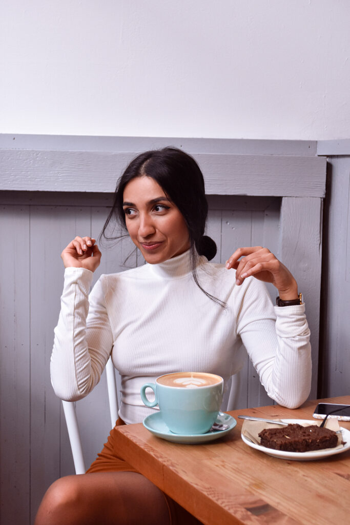 A woman sitting at a wooden table in a café setting, wearing a white turtleneck and smiling off to the side. In front of her is a blue cup of coffee with latte art, and a slice of brownie on a plate, suggesting a cozy, casual moment enjoying a coffee break.