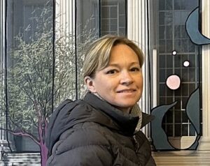 A woman with short blonde hair and a warm smile, wearing a black jacket and a high-collared shirt, stands in front of an abstract mural with a tree motif. The setting gives an urban, contemporary vibe.