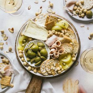 A charcuterie board with a variety of snacks arranged on a round, white plate with a gold rim, on a linen tablecloth. It includes green olives, kiwi slices, brie cheese, rolled ham, crackers, and nuts. There's a glass of white wine to the side and loose nuts scattered around for a casual feel.