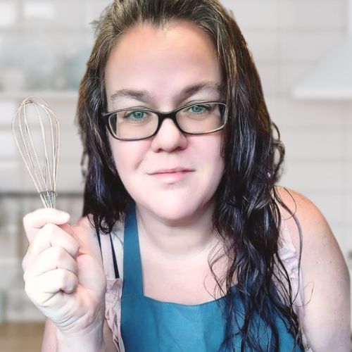 Woman with long, brown, curly hair and glasses holding a ladle and wearing a blue apron.