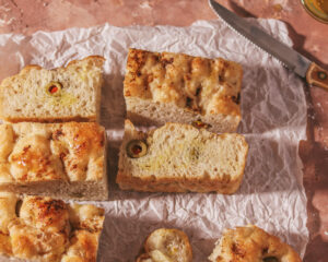 A top-down view of freshly baked focaccia bread with olives and herbs, cut into squares on parchment paper. The bread has a golden brown crust, and the warm tones of the bread are complemented by the rustic table surface, with a kitchen knife to the side suggesting homemade and freshly prepared.