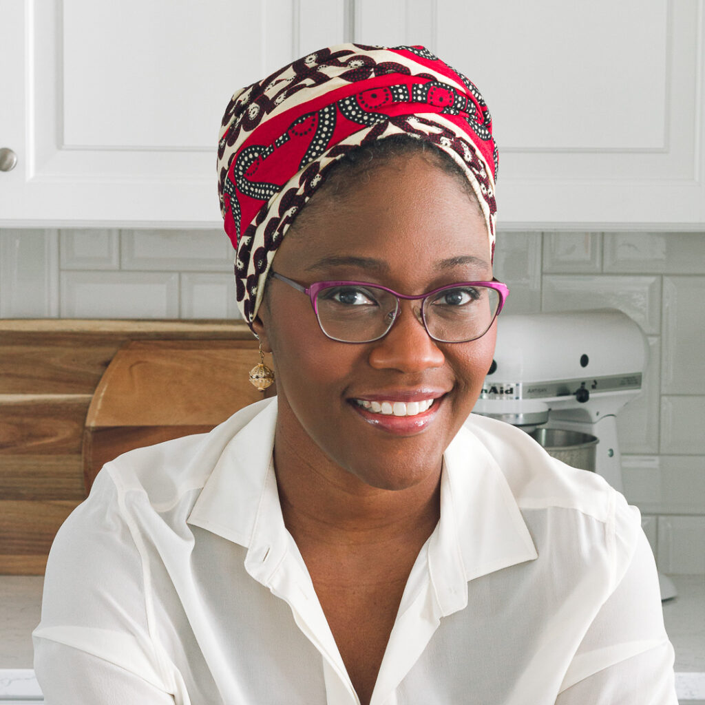 A headshot of a smiling woman wearing a vibrant red and black patterned headscarf and purple glasses. She is dressed in a white blouse, has stud earrings, and is standing in a bright kitchen setting with a white mixer in the background.