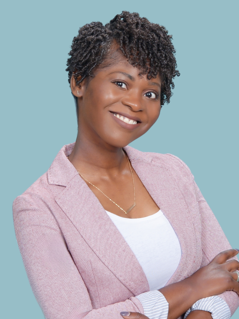 A portrait of a woman with a warm smile, short curly hair, and wearing a pale pink blazer over a white top. Her crossed arms and confident, friendly expression convey professionalism and approachability against a soft blue background.