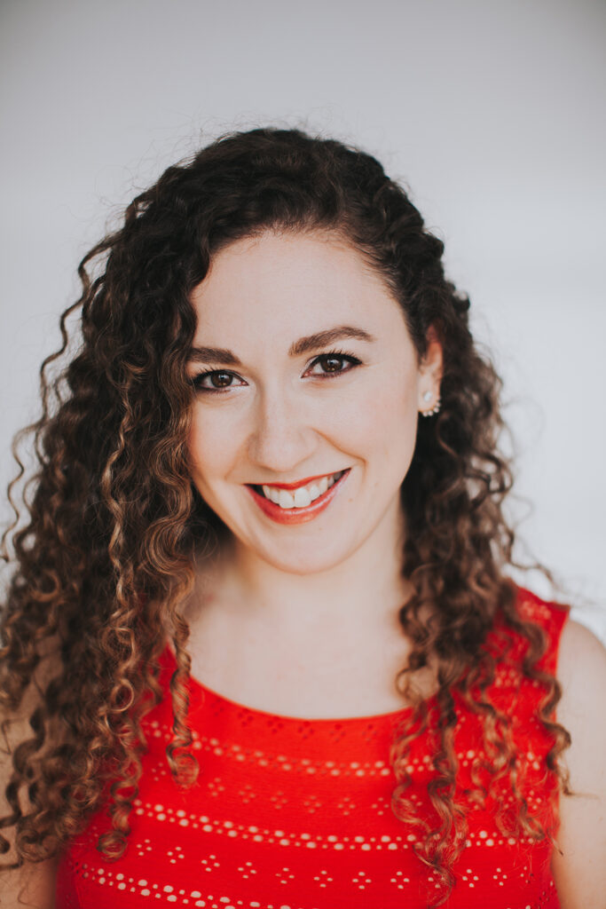 Portrait of a woman with long, dark, curly hair wearing red lipstick and a red shirt. She smiles at the camera.