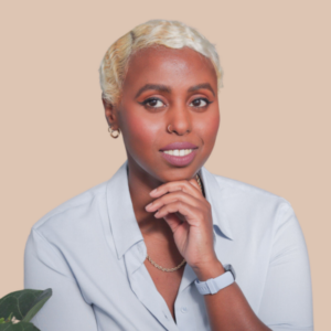 A woman with short platinum blonde hair, wearing a light blue shirt, poses with a hand to her chin. She has a friendly and professional demeanor, and the plain peach background gives the photo a clean and focused feel.