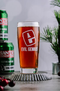 A pint glass filled with amber-colored beer, branded with the words "Evil Genius" in bold red letters. Behind the glass is a can of beer with the text "Santa!! I know him!" and festive decorations including pine branches and cranberries are visible around the scene.