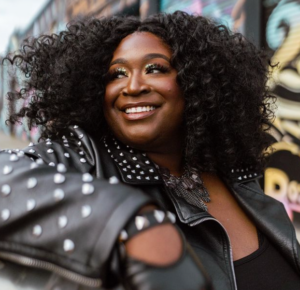 A joyous woman with a radiant smile, wearing a stylish black leather jacket adorned with studs. She has voluminous curly black hair and her makeup features a subtle shimmer. She appears in an urban setting with graffiti art in the background, conveying a sense of vibrant street style.