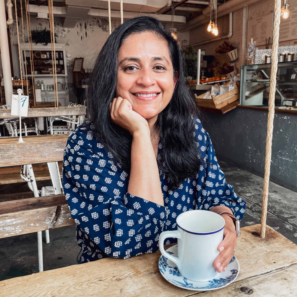 A woman with mid-length dark hair sits in a cafe with wooden tables. She holds a white ceramic mug of coffee in one hand, wears a polka dot blue blouse, and rests her hand on her chin.